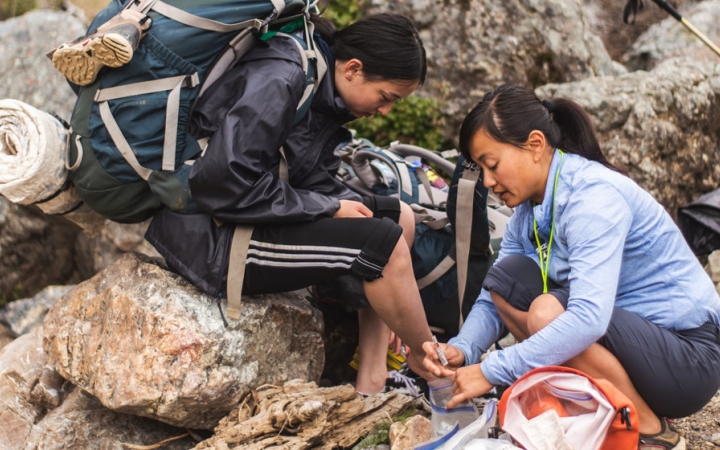 a person wearing backpacking gear sits on a rock while another person helps them on an outward bound course for bipoc